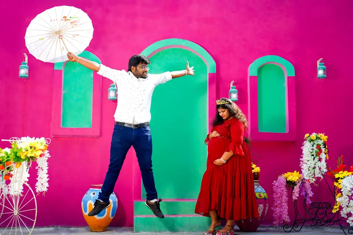 Man jumps joyfully while mom-to-be watches in red gown at green-pink wall