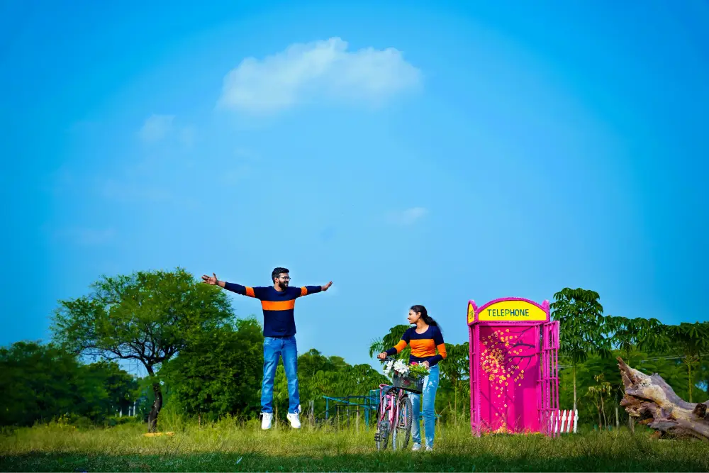Couple by pink telephone booth and bicycle in open field under blue sky – pre wedding Hyderabad